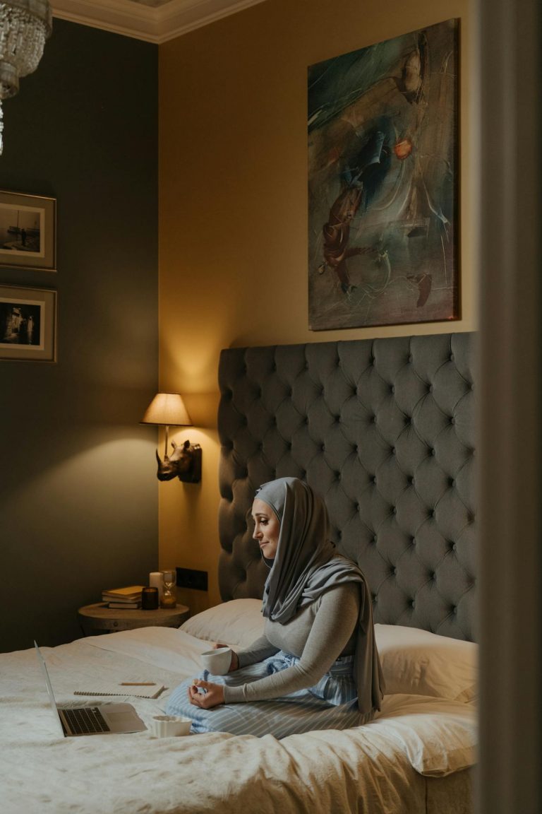 A woman in a hijab sits with her laptop in a warm, stylish bedroom, drinking coffee and browsing online.