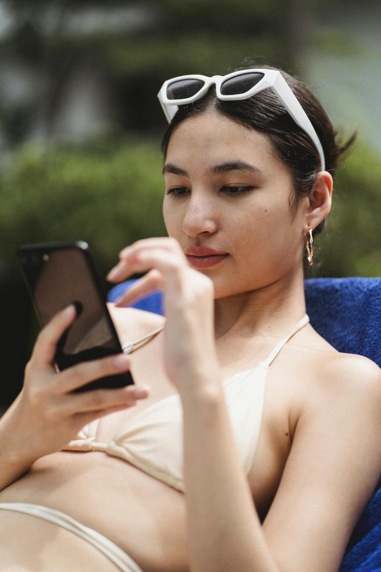 Concentrated young ethnic female tourist in bikini reading news on mobile phone while chilling in deckchair in yard of hotel